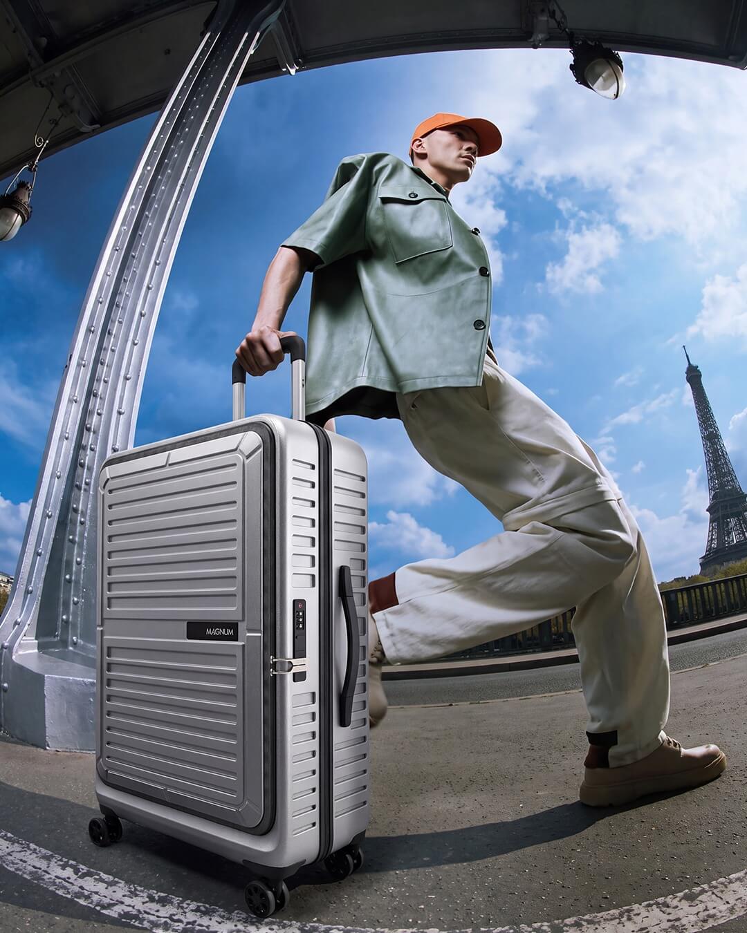 A man in a green shirt, beige pants, and an orange cap pulls a silver ribbed hard-shell suitcase under a bridge with the Eiffel Tower in the background.